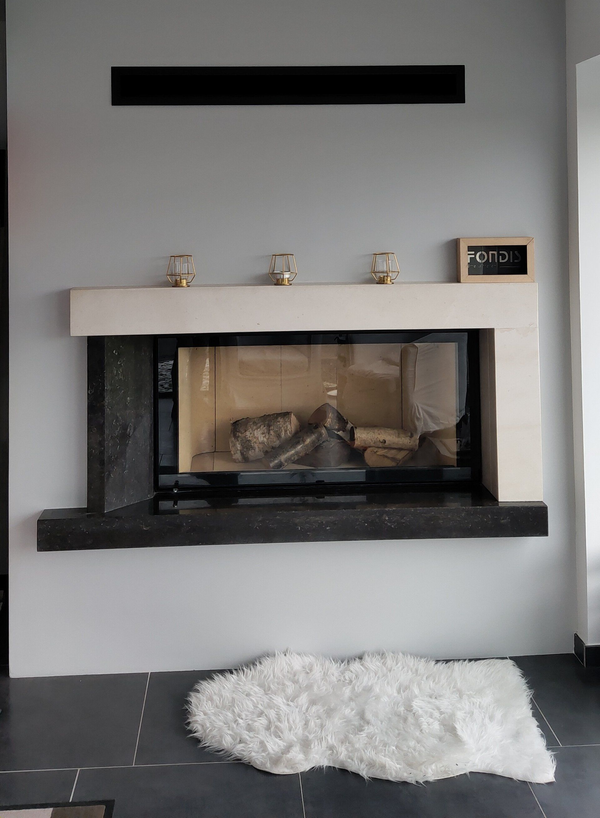 Cheminée dans un salon, avec un manteau noir et blanc, une grille d'aération murale et un tapis blanc sur un sol foncé.