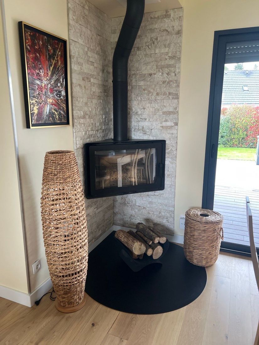 Coin cheminée moderne avec poêle suspendu noir, bois de chauffage, paniers tressés et porte vitrée donnant sur le jardin.