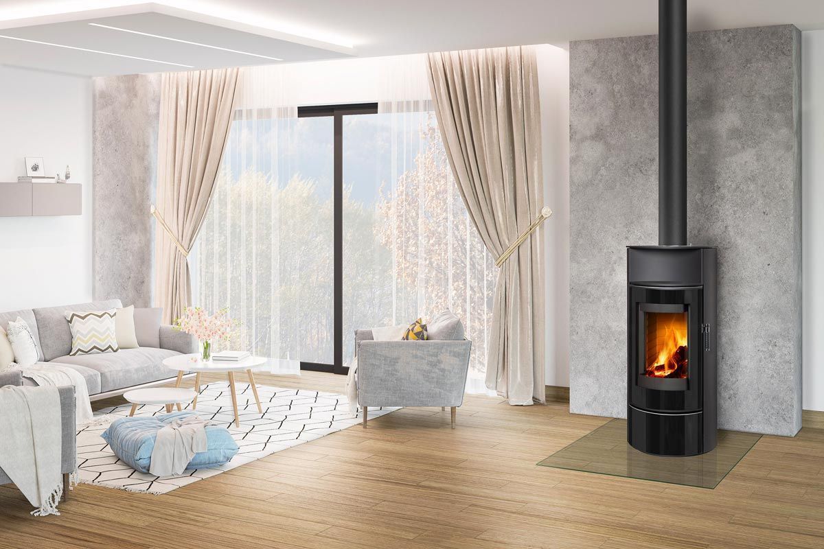 Salon moderne et lumineux avec une grande fenêtre, un canapé à motifs et un poêle à bois noir sur la droite.