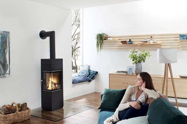 Salon moderne avec un poêle à bois noir, un canapé bleu canard et une femme se détendant près d'une fenêtre lumineuse.