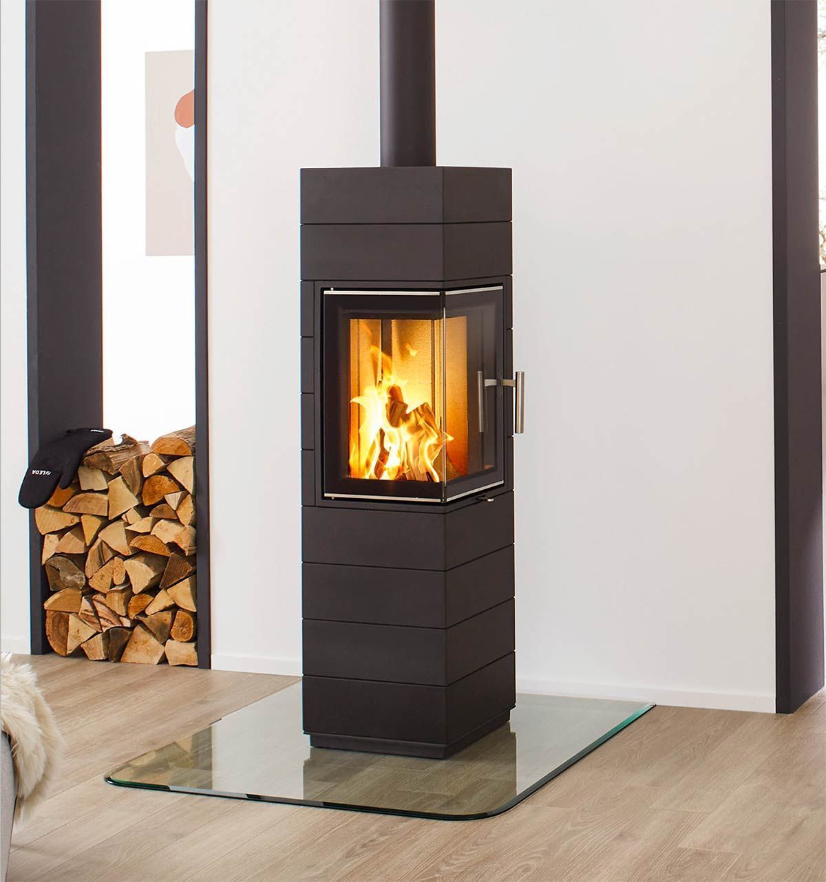 Poêle à bois noir avec flammes visibles, à côté d'une pile de bois de chauffage sur un foyer vitré dans une pièce lumineuse.