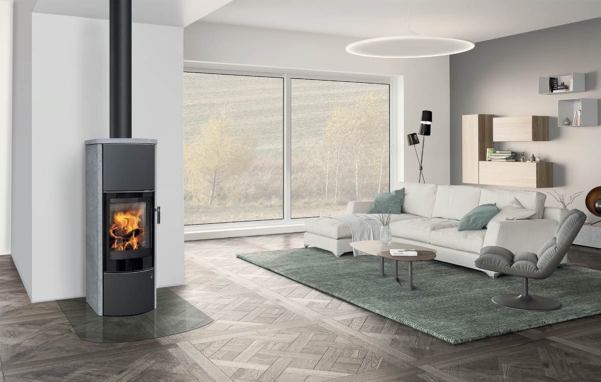 Salon moderne avec un poêle à bois indépendant allumé, de grandes fenêtres, un canapé gris et un tapis vert.