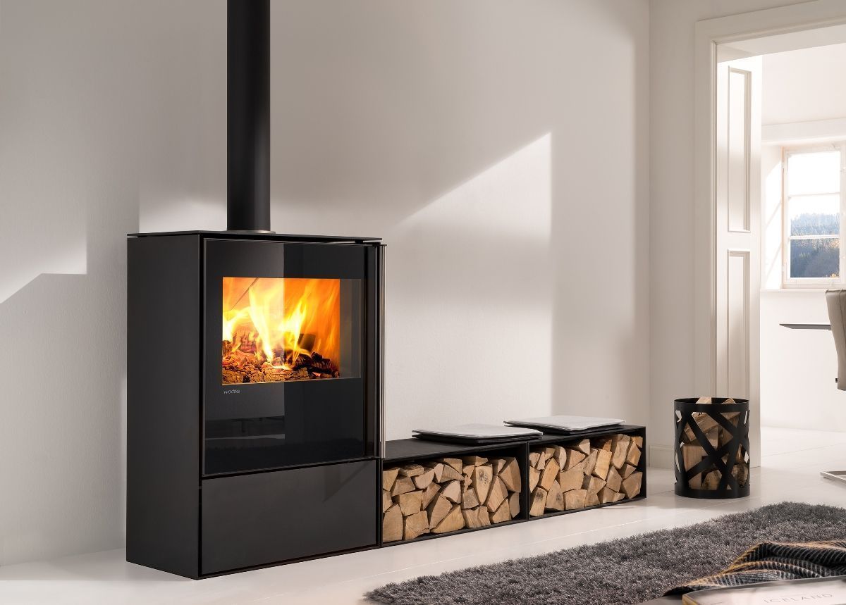 Poêle à bois noir moderne avec un feu allumé dans un salon lumineux, à côté d'une pile de bois de chauffage.