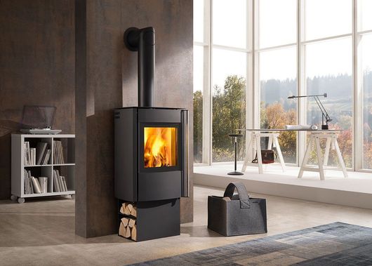 Salon moderne avec un poêle à bois noir, un feu qui crépite, de grandes fenêtres et du bois de chauffage empilé à proximité.