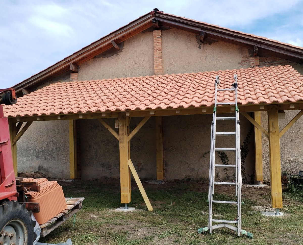 Un nouvel abri de voiture en tuiles de terre cuite est en cours de construction contre un bâtiment en briques beiges.