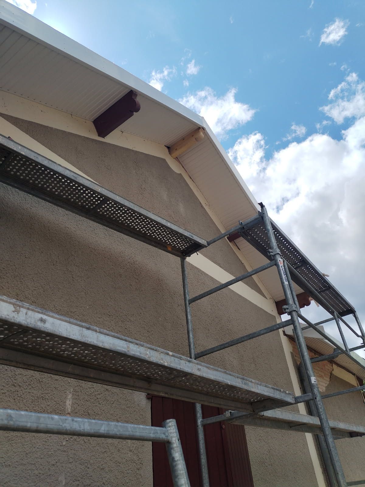 Échafaudage à côté d'un bâtiment avec chevrons de toit exposés et soffite blanc ; ciel bleu au-dessus.