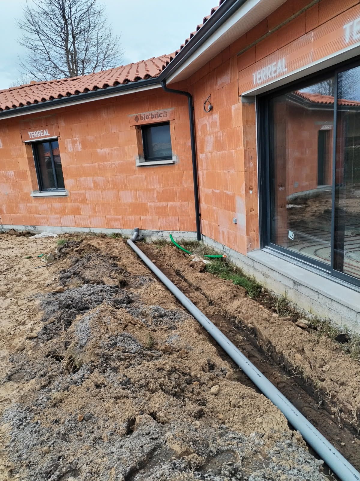 Extérieur d'un bâtiment en construction avec tranchée et canalisation. Murs de briques, fenêtres noires.