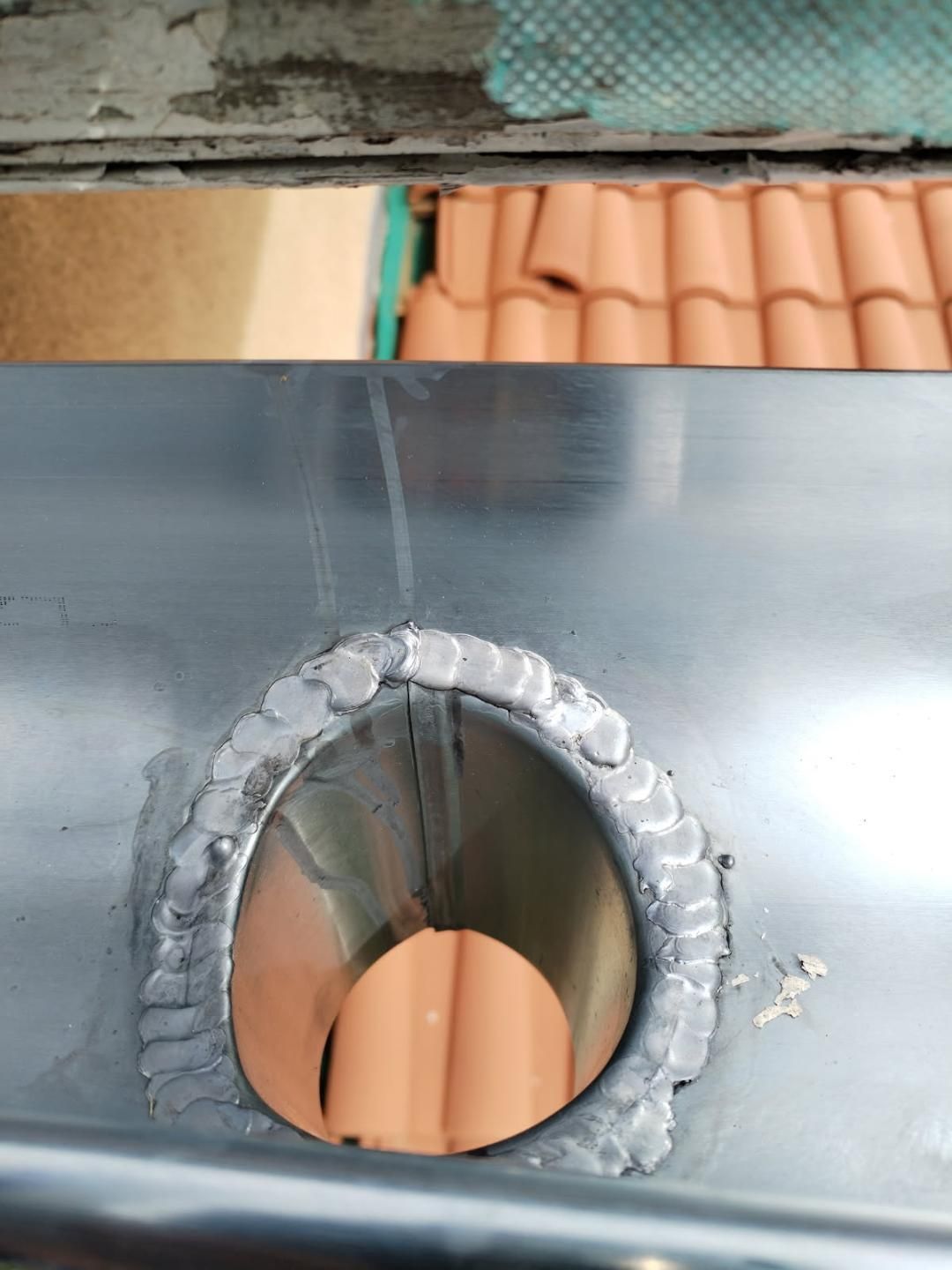 Tuyau en acier soudé à une surface métallique, probablement une gouttière. Toit orange visible en arrière-plan.
