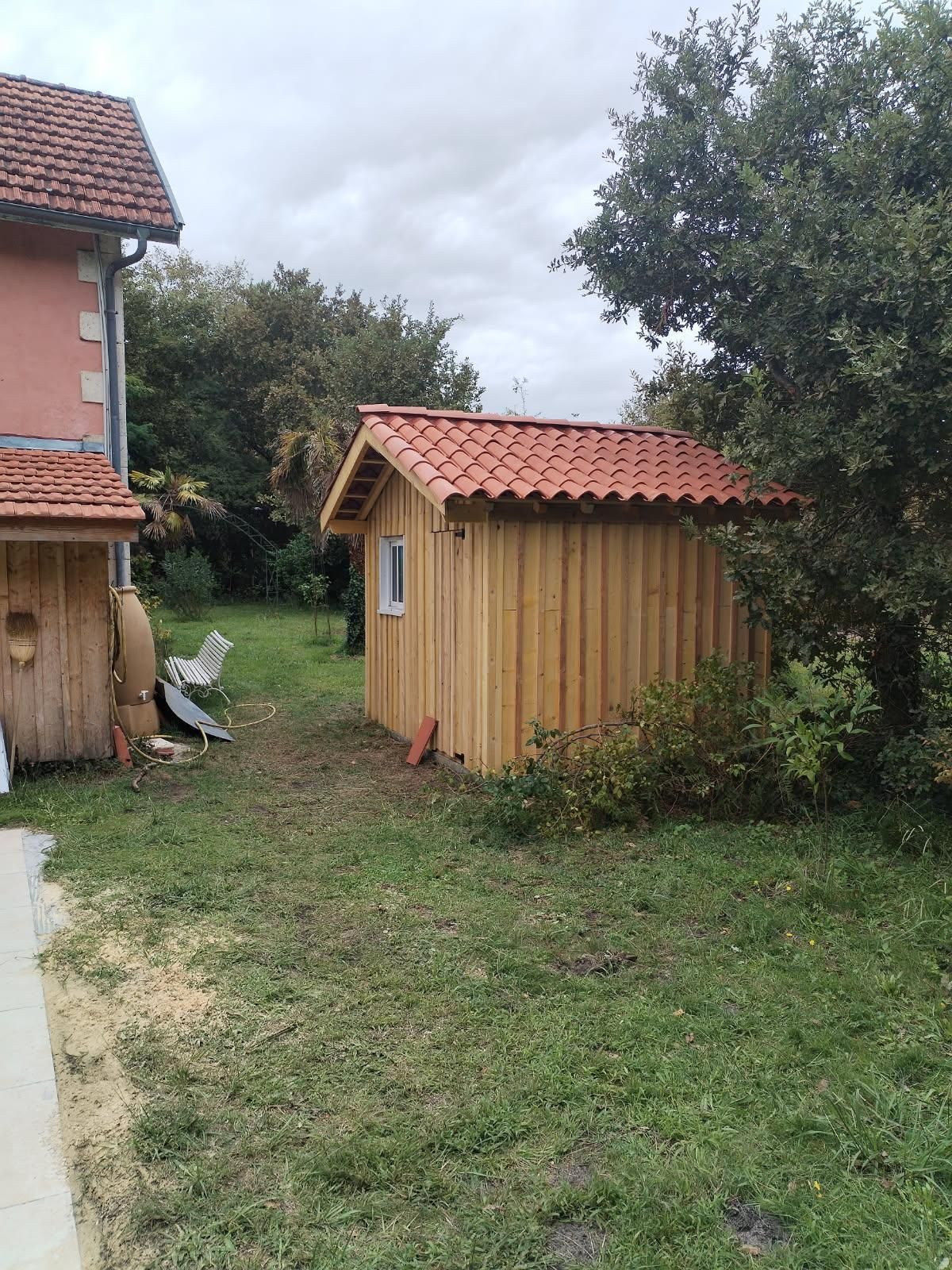 Hangar en bois avec toit en tuiles rouges, situé dans une cour arrière herbeuse avec des arbres et un bâtiment rose.
