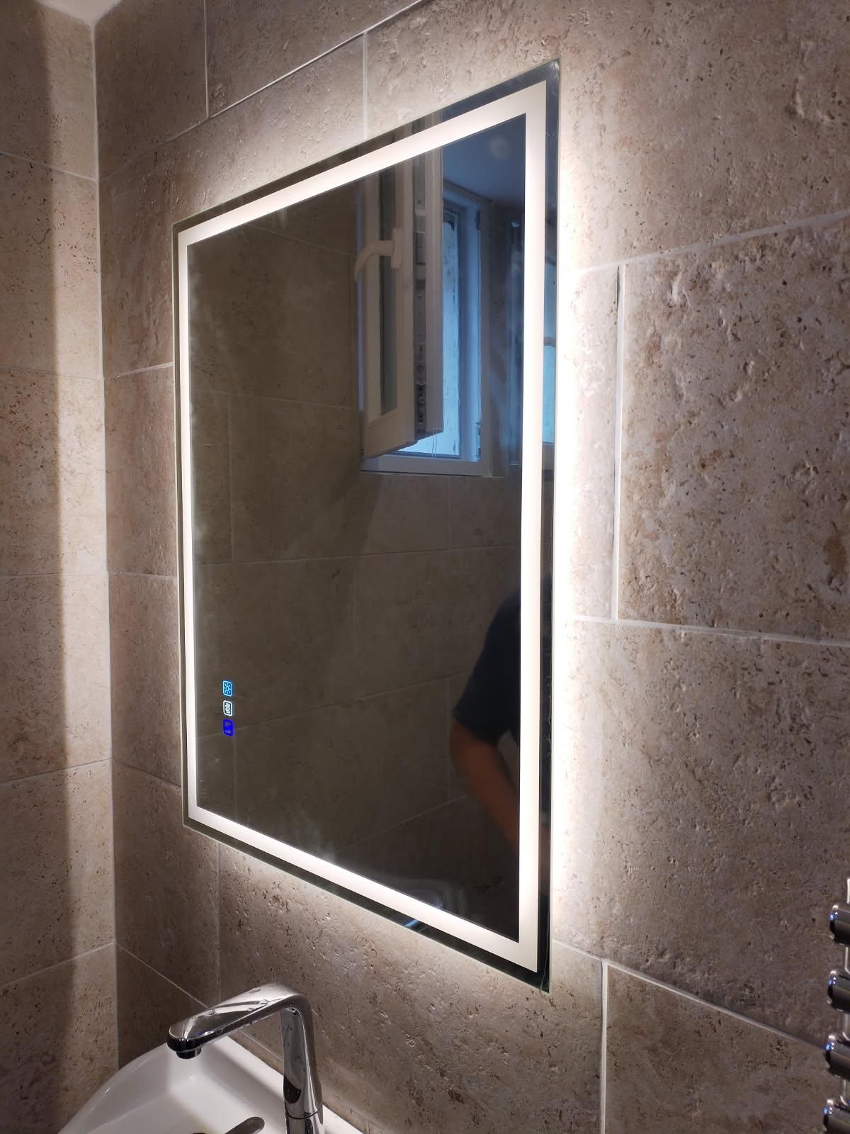 Miroir de salle de bains rectangulaire éclairé, adossé à un mur carrelé beige clair. Robinetterie chromée.