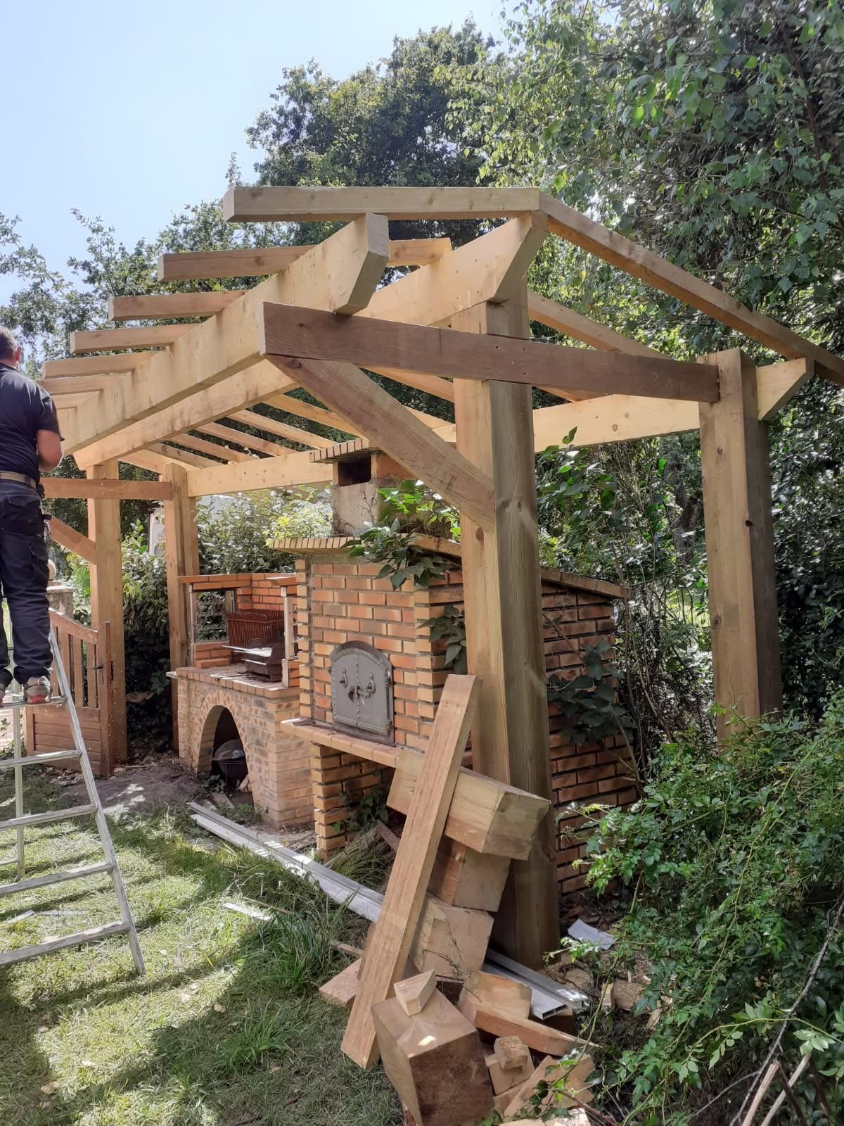 Un homme construit une pergola en bois sur un four extérieur en briques dans un jardin ensoleillé.