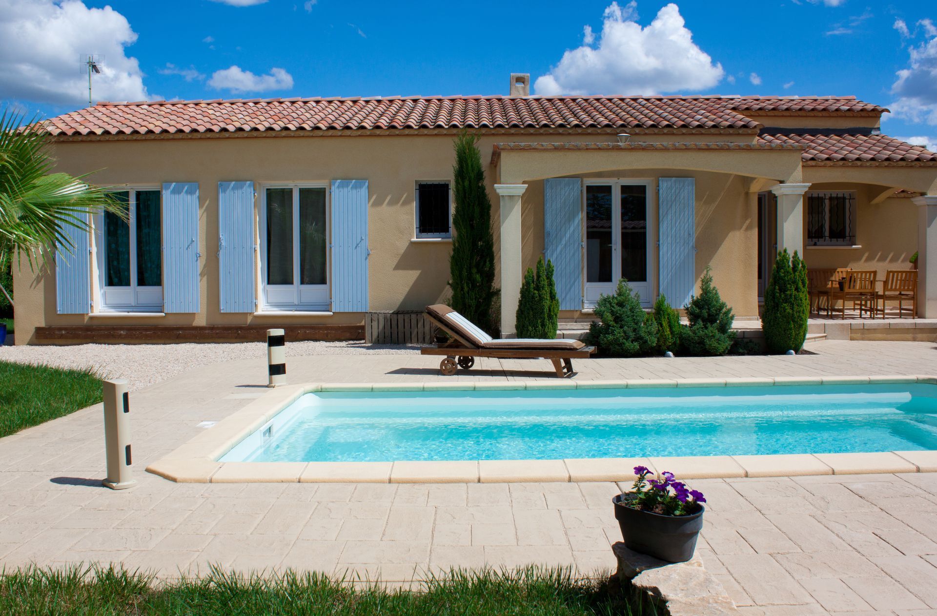 Maison avec volets bleu clair, piscine et chaise longue par une journée ensoleillée.