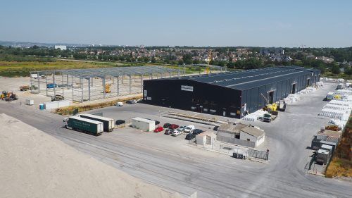 Chantier de construction avec deux bâtiments : un entrepôt noir terminé et une structure à ossature d'acier en construction.