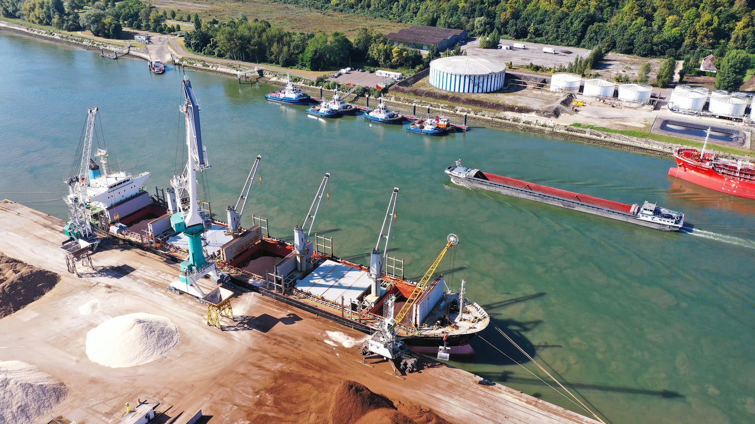 Navire en cours de déchargement près de la rive du fleuve, avec bateaux de soutien, rivière et structures industrielles visibles.