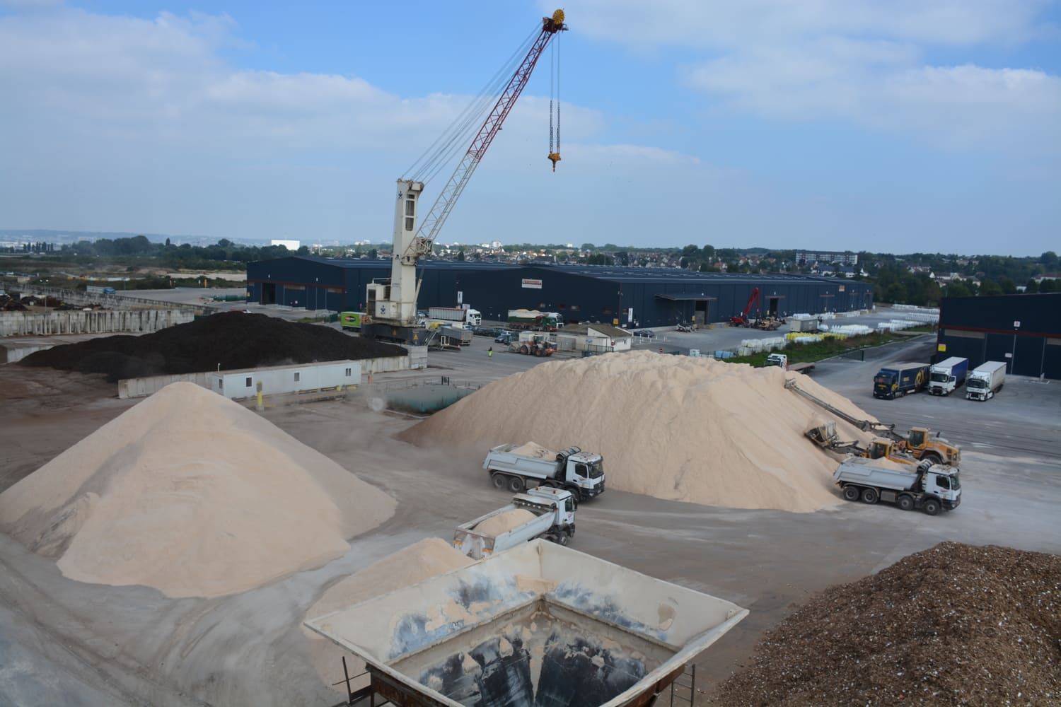 Chantier de construction avec de gros tas de sable, une grue et des camions ; bâtiment moderne en arrière-plan sous un ciel nuageux.