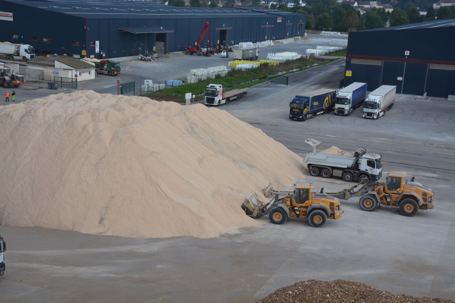 Deux chargeuses jaunes extraient du sable d'un gros tas ; des camions attendent à proximité.