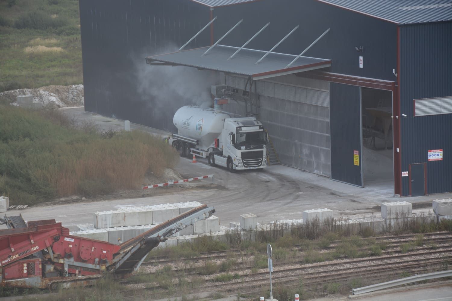 Un camion-citerne entre dans un entrepôt, soulevant des volutes de poussière blanche. Bâtiment gris, blocs de béton et machines en vue.