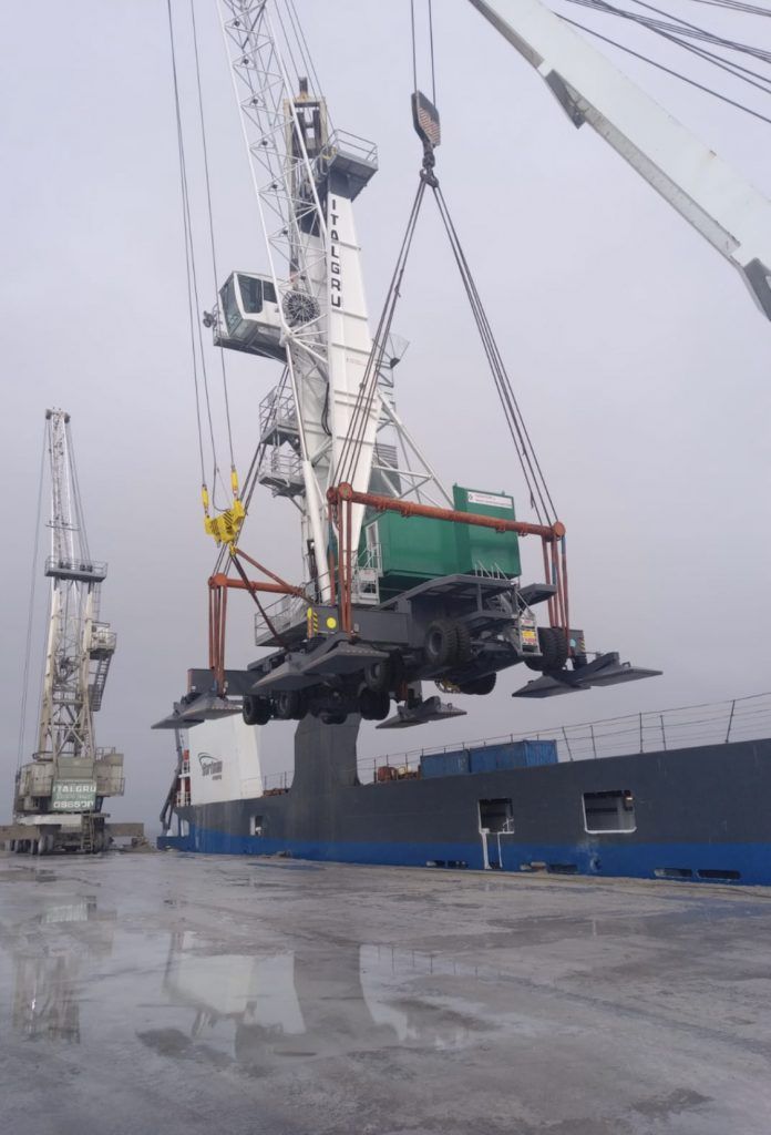 Une grande grue soulevant une machine sur un navire dans un port, ciel couvert.