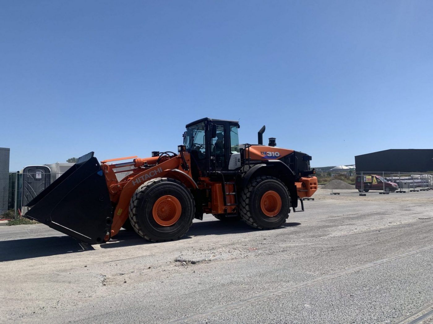 Chargeuse sur pneus orange garée sur un terrain en gravier sous un ciel bleu clair.
