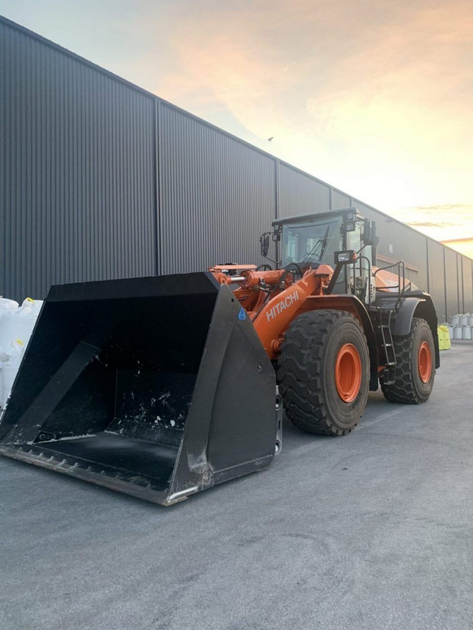 Chargeuse sur pneus Hitachi orange devant un bâtiment sombre au coucher du soleil.