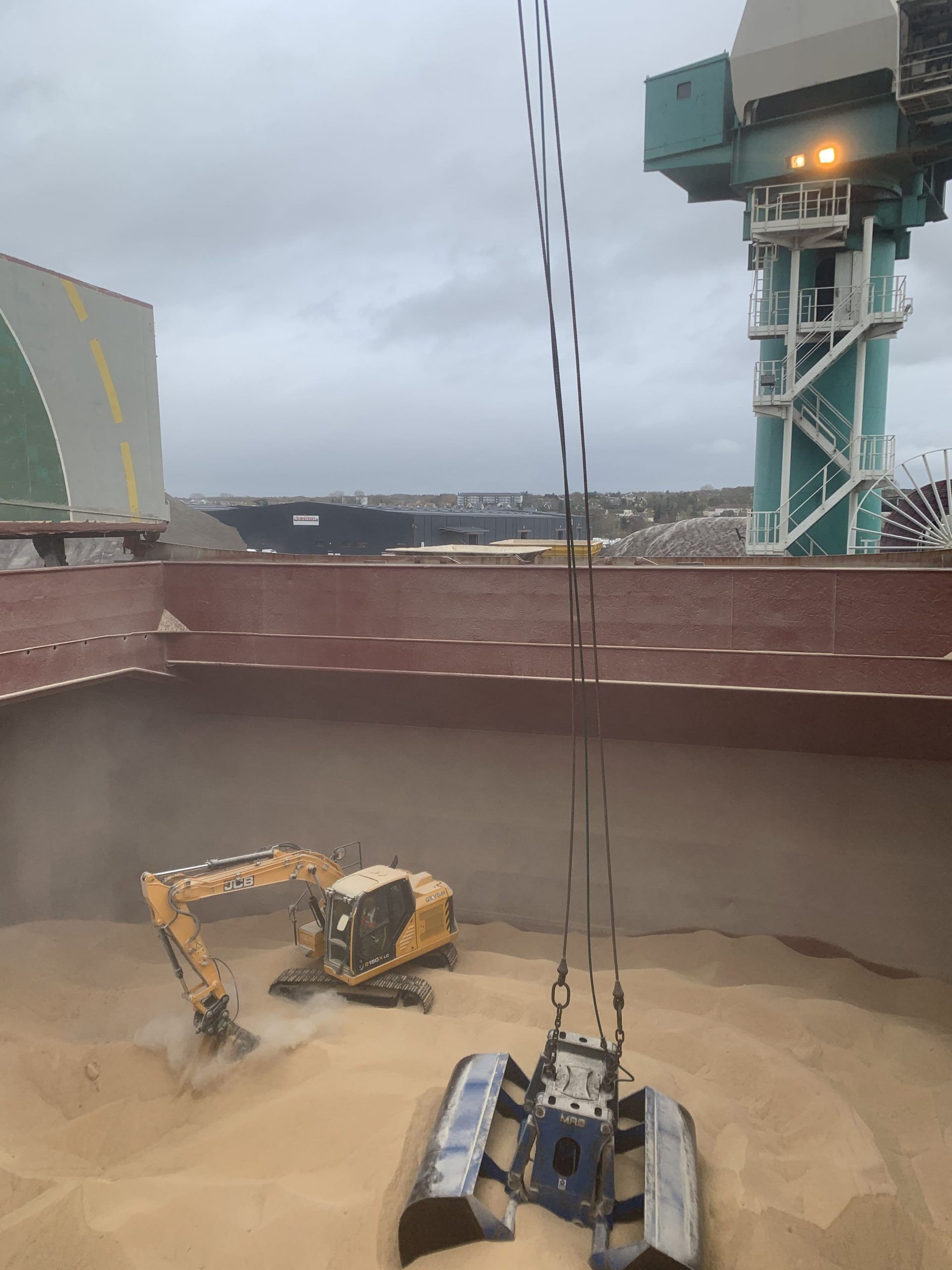 Excavateur et grue à benne preneuse déchargeant du grain à l'intérieur d'un cargo, en milieu industriel.
