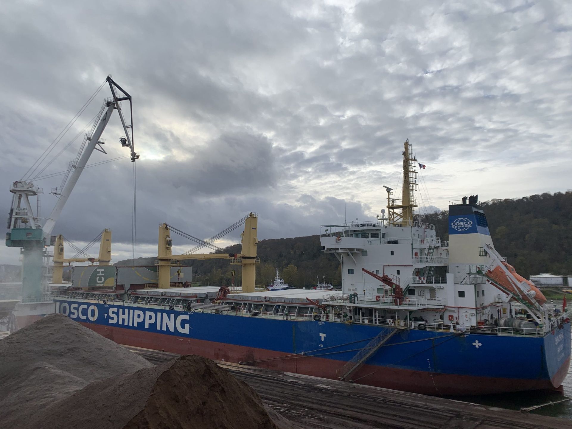 Un cargo est en cours de chargement dans un port ; une grue charge la cargaison sous un ciel nuageux.