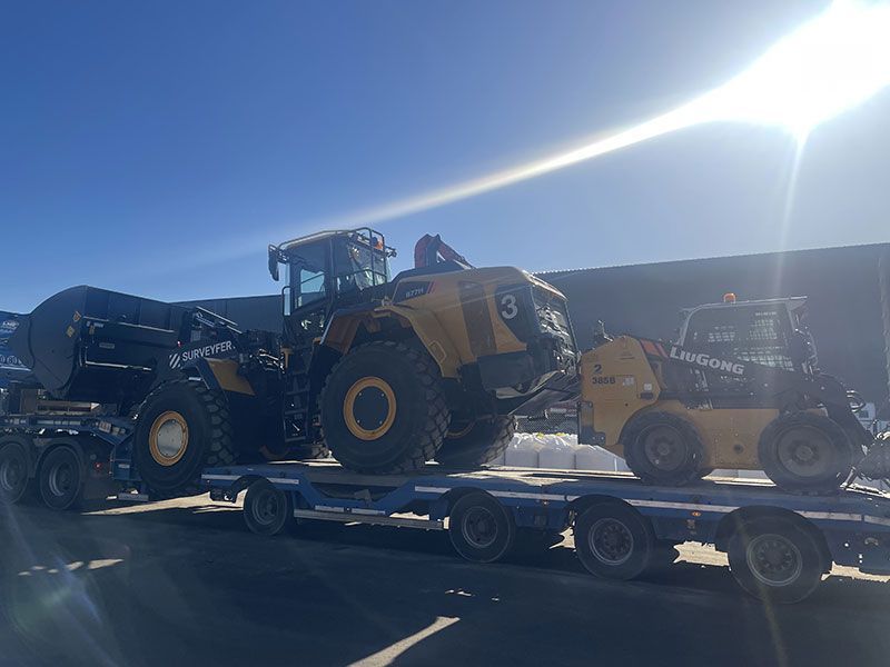 Trois véhicules de construction chargés sur une remorque à plateau sous un ciel lumineux et ensoleillé.