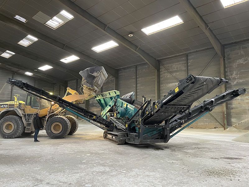 Un chargeur frontal jaune et un concasseur de roche vert à l'intérieur d'un grand entrepôt.