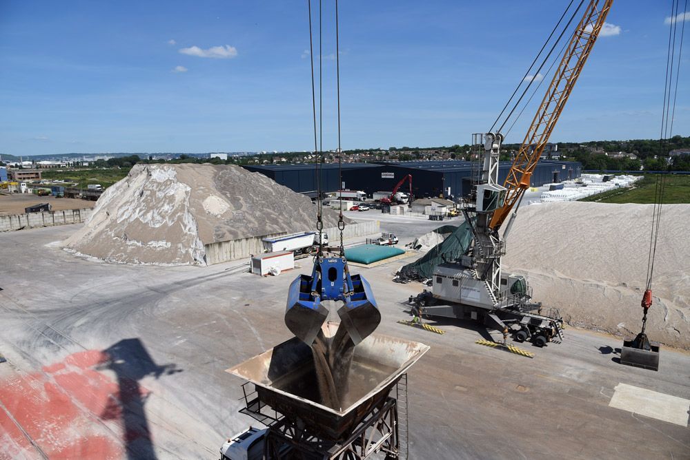 Grue chargeant des matériaux dans une trémie sur un chantier de construction avec des tas de gravier en arrière-plan.