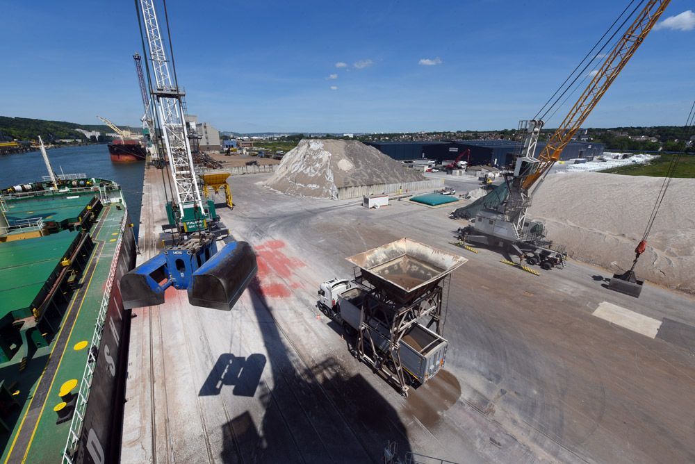 Un port avec des grues chargeant un navire. Des piles de matériaux et une machine dans le port.