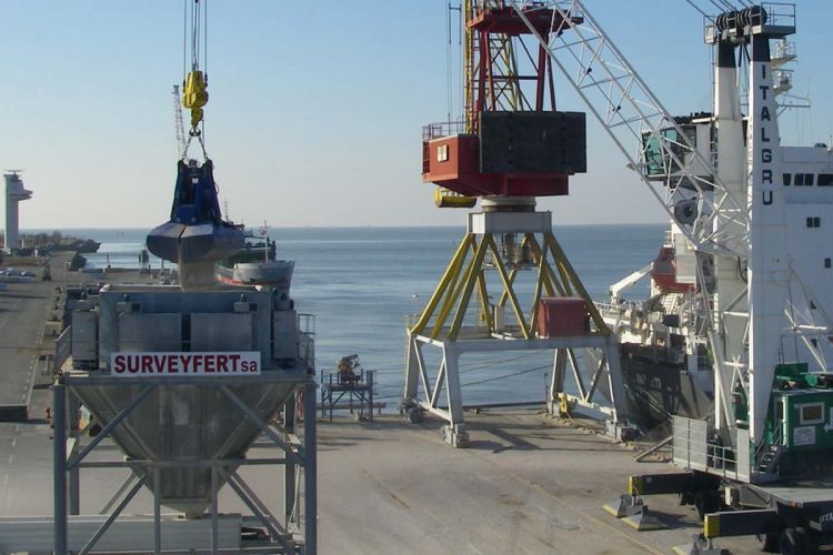Grue déchargeant de l'engrais d'un navire. Structure métallique avec benne, conteneur et grue rouge à quai.