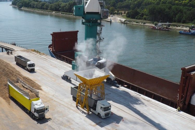 Chargement de céréales depuis une grue vers un cargo dans un port fluvial, chargement de camions à proximité.