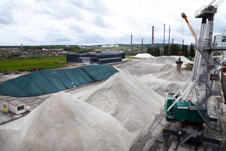 Scène portuaire : tas de matériaux de couleur claire, grue, bâche verte, bâtiments, cheminées, ciel nuageux.