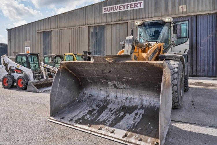 Machinerie lourde devant un entrepôt avec le panneau « SURVEYFERT ». Une grande chargeuse frontale domine la vue.