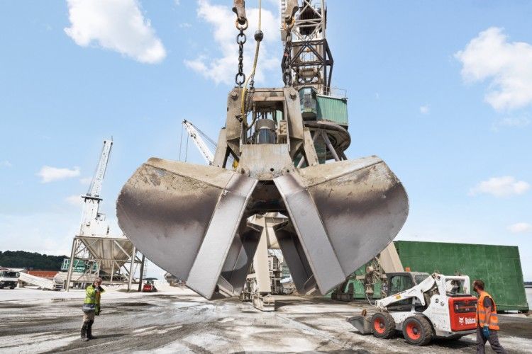 Grue à grande pince déchargeant du matériel dans un port. Deux ouvriers portant des gilets de sécurité se trouvent à proximité. Ciel bleu.