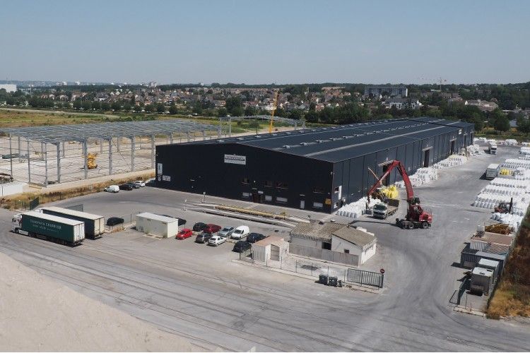 Vue aérienne d'un grand bâtiment industriel noir avec des camions, des voitures et des travaux de construction à proximité ; journée ensoleillée.
