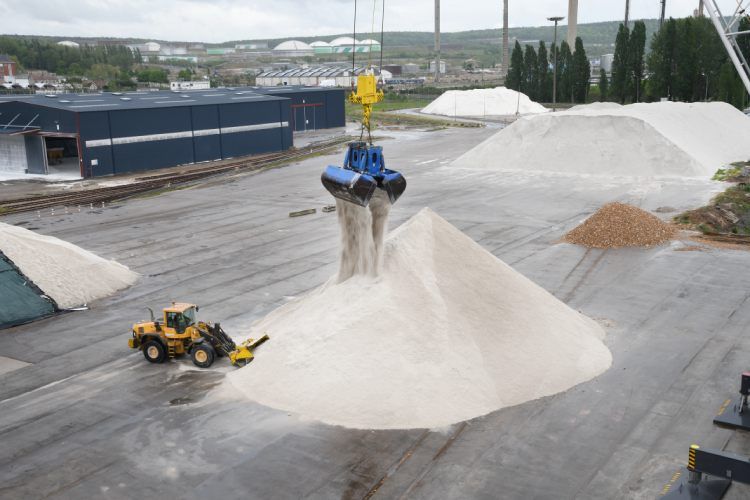 Une grande grue dépose du matériau blanc sur un tas ; une chargeuse jaune est à proximité. Environnement industriel.