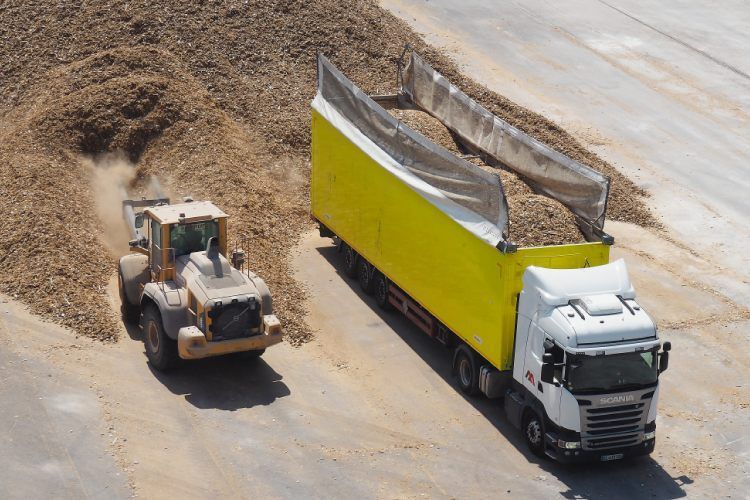 Un camion jaune chargé de copeaux de bois par un chargeur frontal ; à l'extérieur sur une surface en béton.
