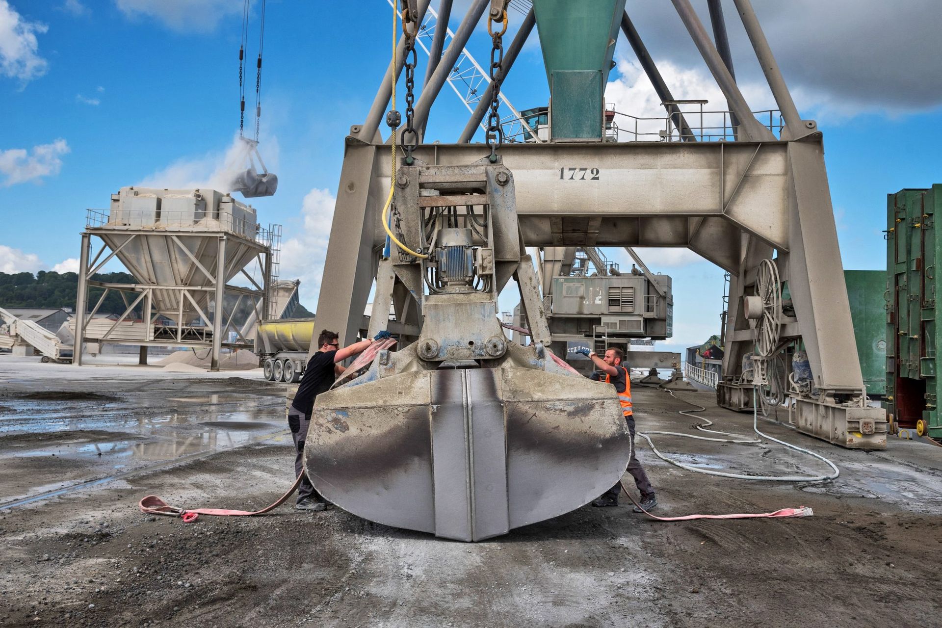 Deux ouvriers manœuvrant une grande benne preneuse sur un site industriel.