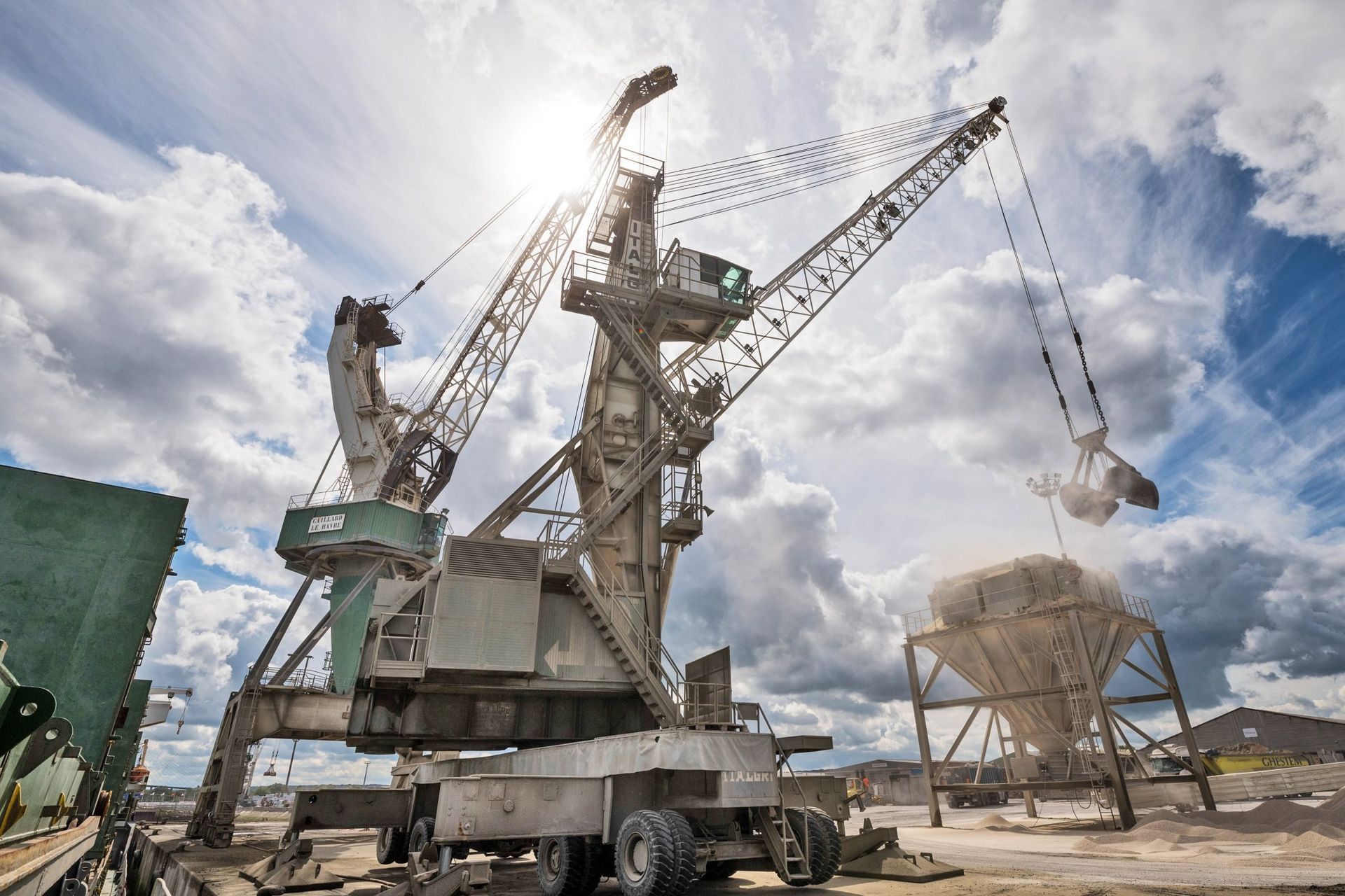 Une grande grue industrielle soulève du matériel contre un ciel ensoleillé et nuageux.