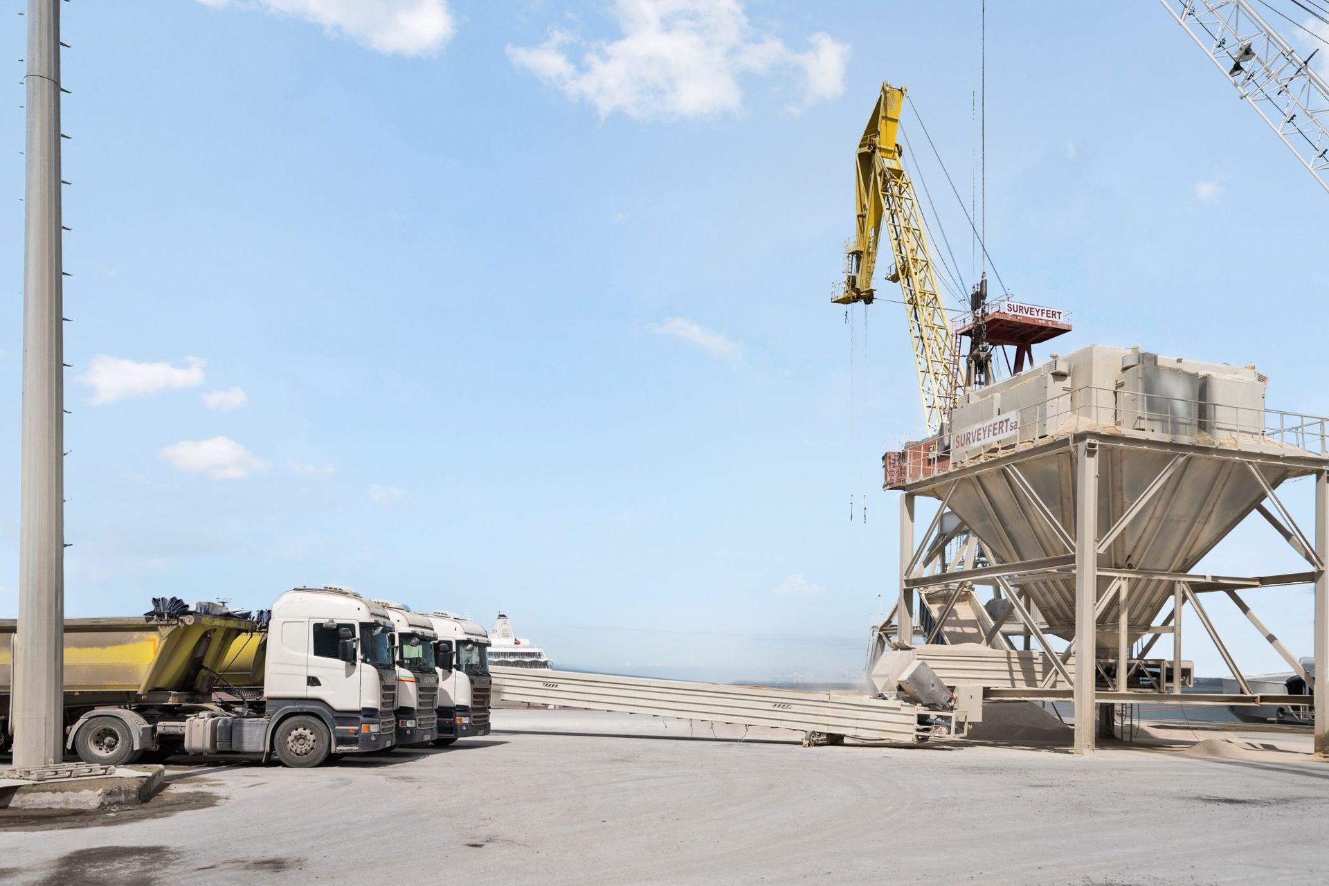 Des camions alignés sur un chantier de construction avec une grue, un silo et un ciel bleu.