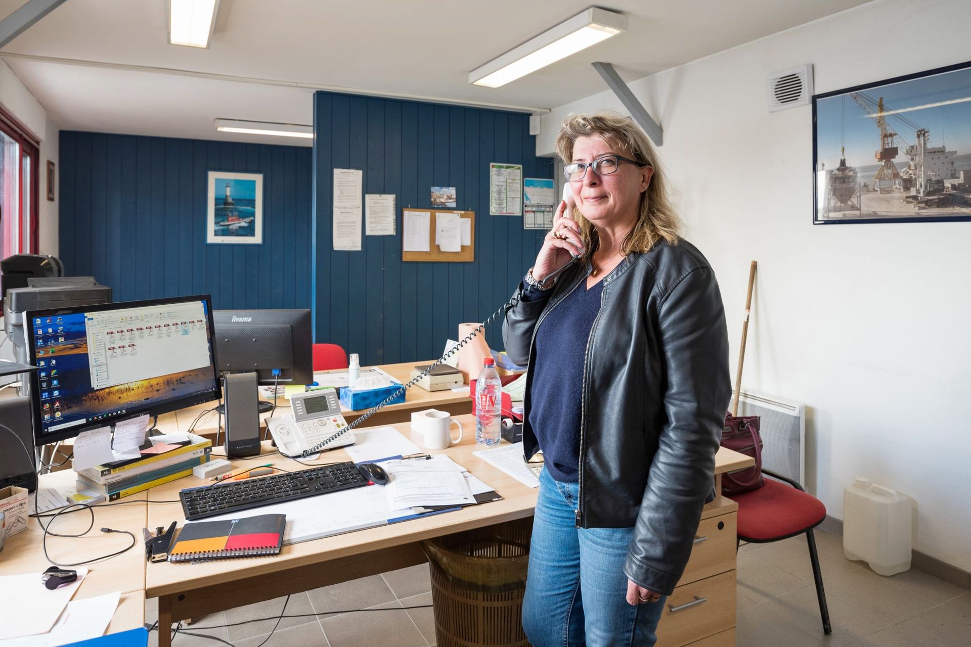 Une femme au bureau, appuyée sur son bureau, téléphone. Elle porte une veste et un jean. Le bureau est équipé d'un ordinateur.
