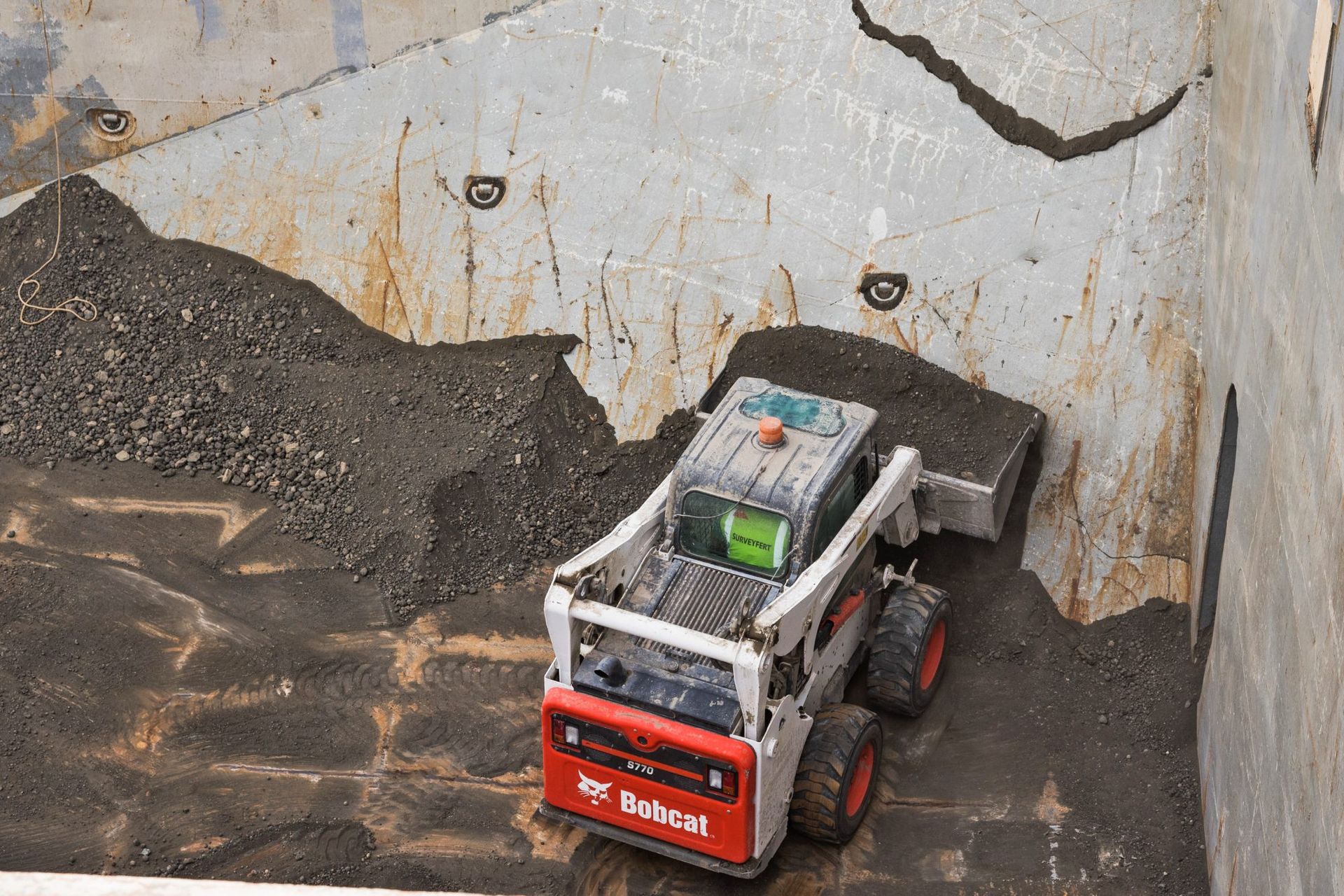 Une petite chargeuse compacte Bobcat ramasse du gravier foncé dans un conteneur à parois métalliques.