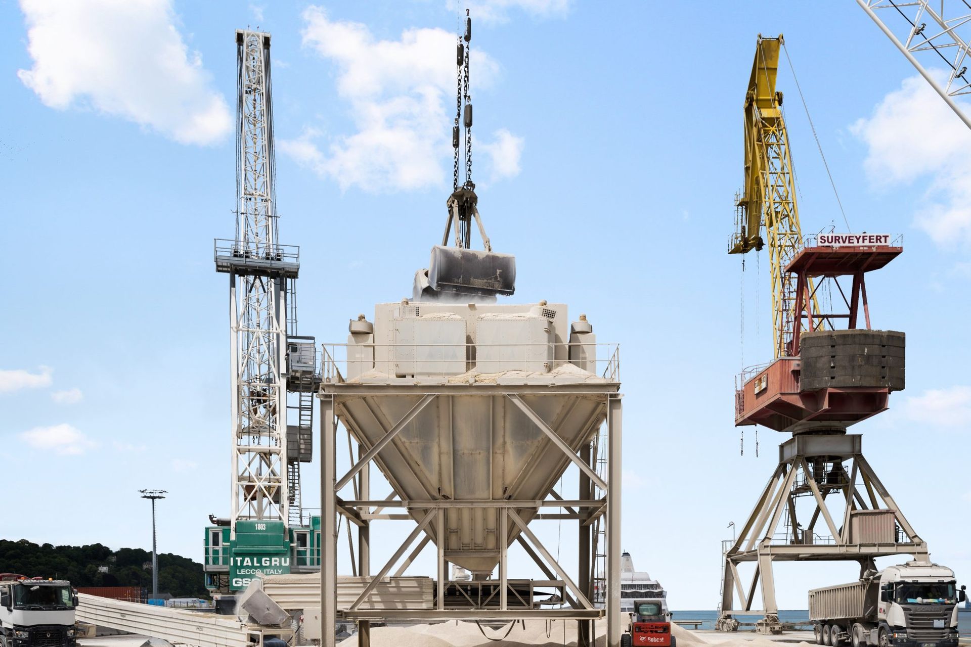 Grues chargeant une grande trémie dans un port, ciel bleu au-dessus.