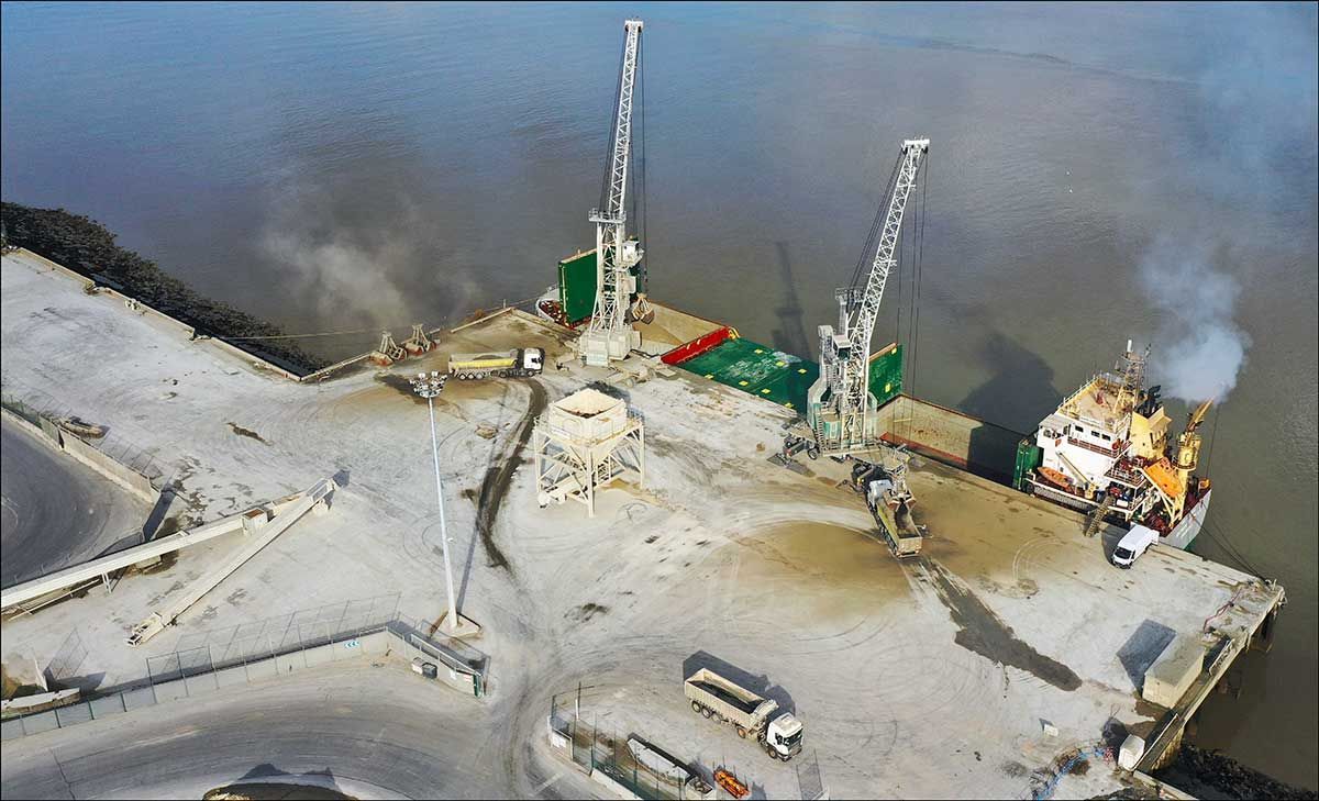 Vue aérienne d'un port avec des grues chargeant des marchandises sur un navire. Quai gris, sable beige et eau calme.