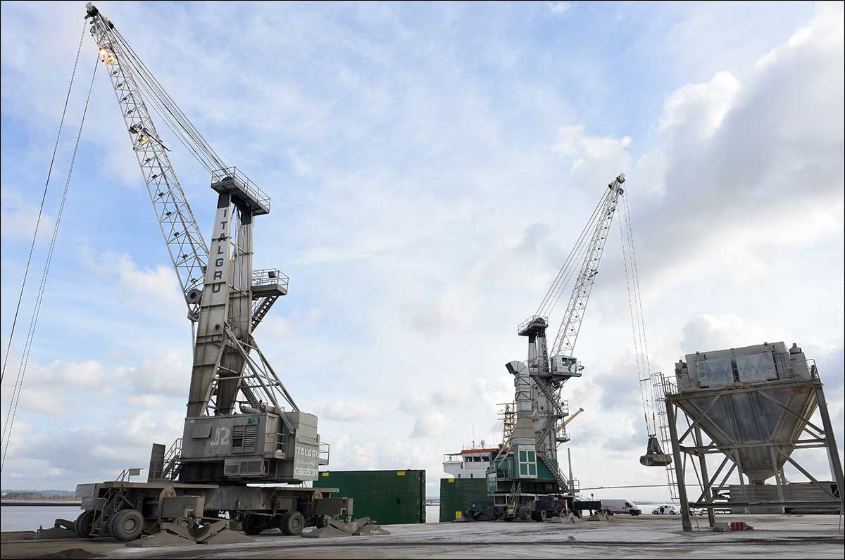 Deux grandes grues dans un port chargeant des matériaux, sous un ciel nuageux.