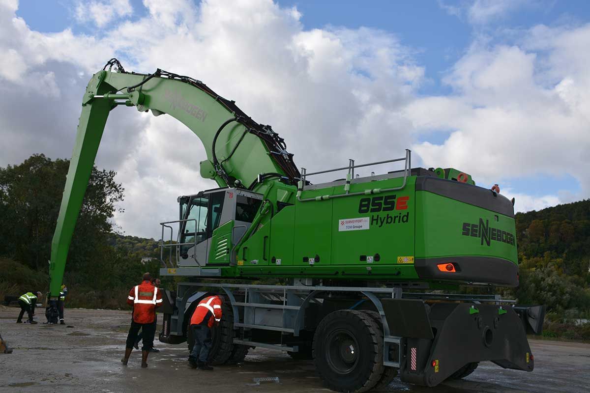 Excavatrice hybride verte sur roues sur un chantier de construction ; deux ouvriers à proximité ; ciel nuageux.