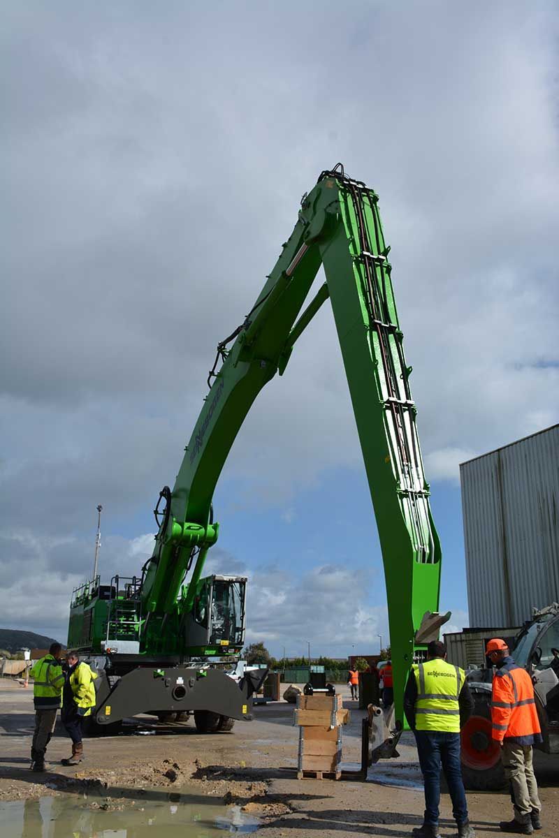 Une excavatrice verte s'élève haut, travaillant sur un chantier avec des ouvriers à proximité. Ciel nuageux.