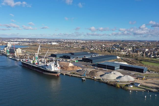 Cargo amarré dans un port, à côté de bâtiments industriels et de piles de matériaux. Ciel bleu et ville en arrière-plan.