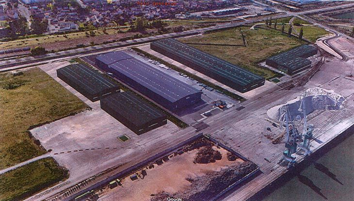 Vue aérienne de bâtiments industriels aux toits longs et sombres, à proximité d'un front de mer et de voies ferrées.